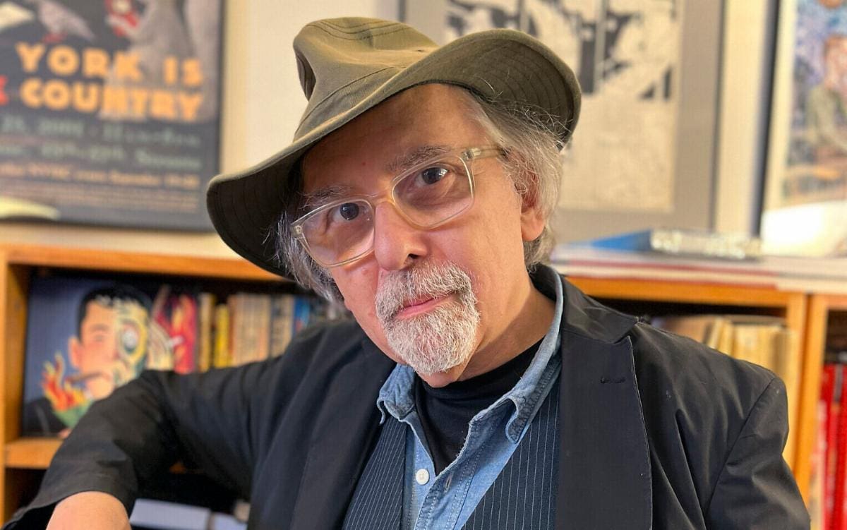 A man in a hat sitting in front of a bookshelf.